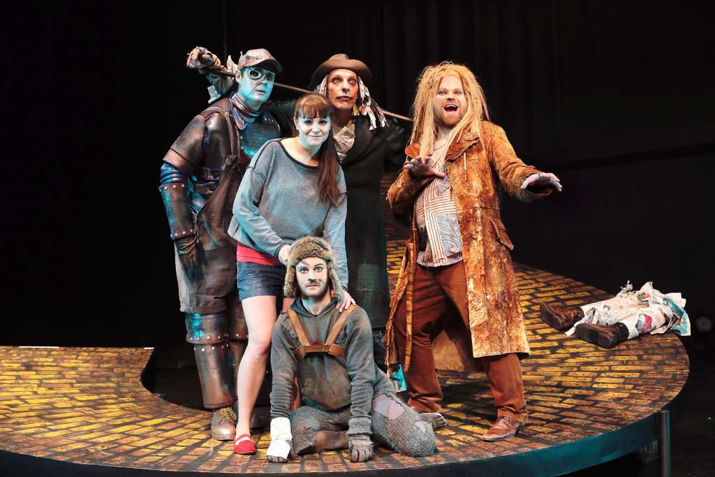 Viisi näyttelijää teatterin lavalla rykelmässä, osa katsoo hymyillen kameraan ja osa kameran ohi.