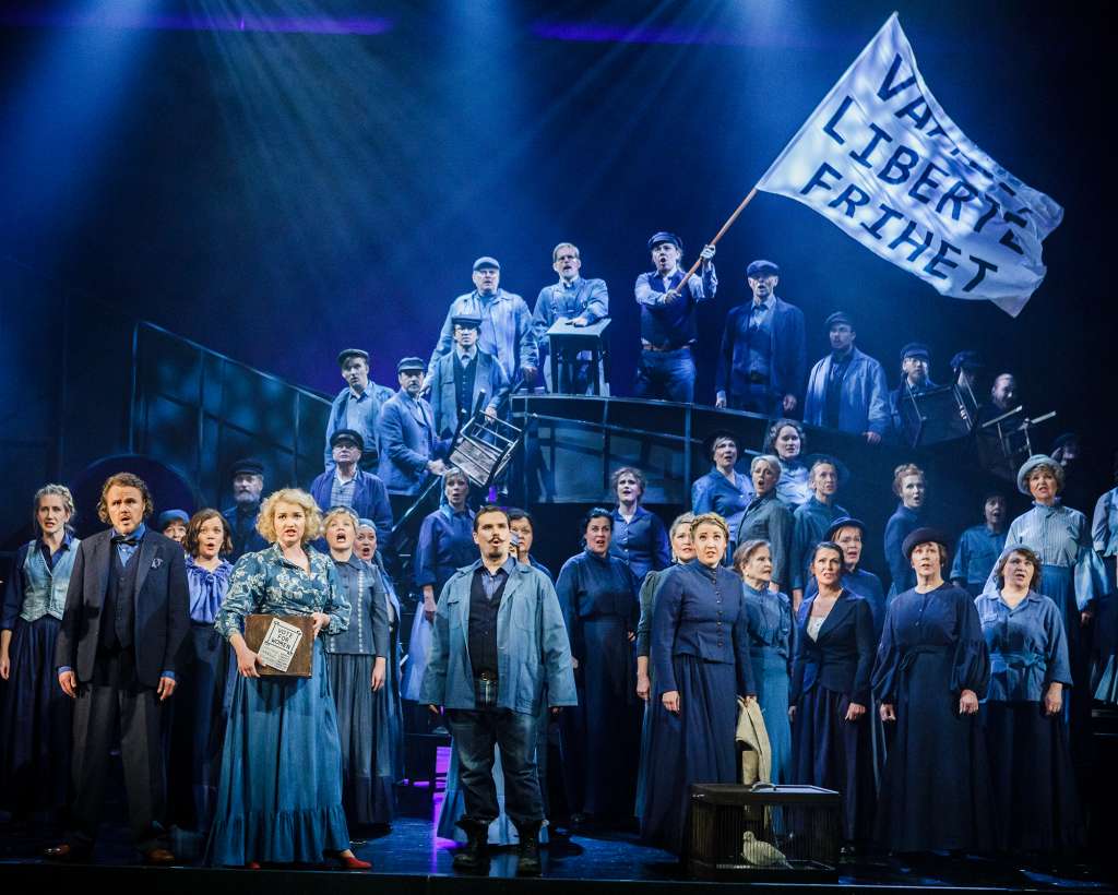 Sinisessä valossa suuri joukko näyttelijöitä ja laulajia katsovat kameran ohi ja laulavat. Osa seisoo korokkeella, jossa mies pitelee lippua, jossa lukee vapaus, liberty, frihet.