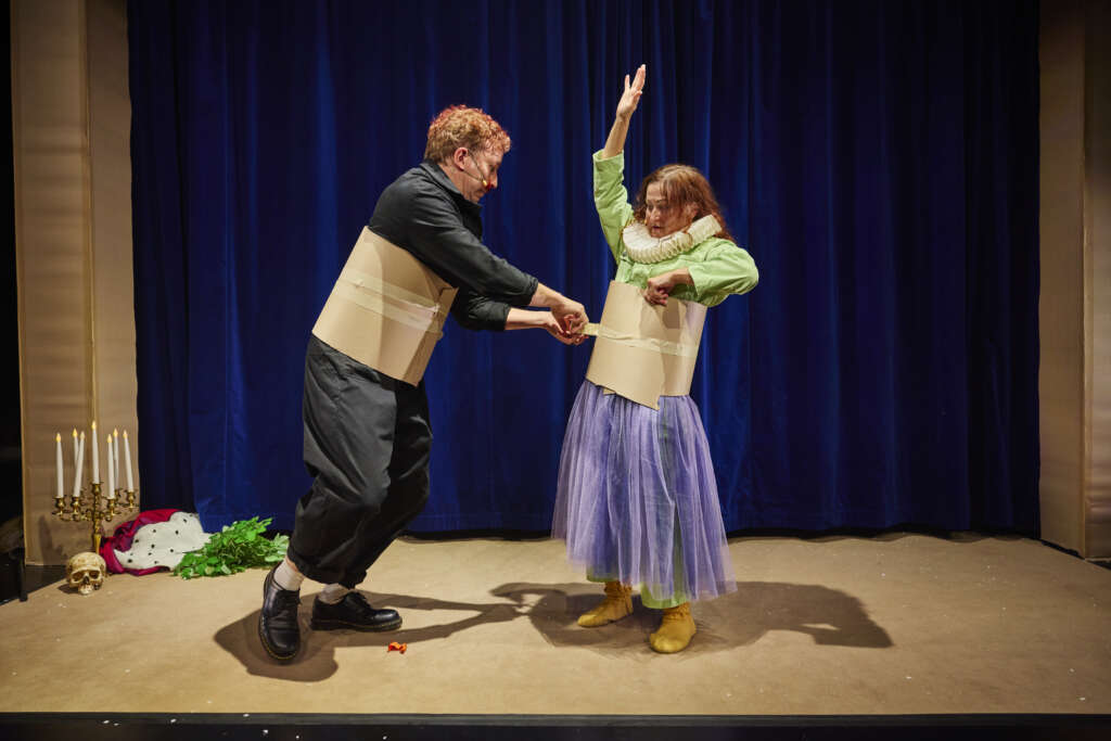 Tummaan asuun pukeutunut mies kiertää teippiä värikkääseen asuun pukeutuneen naisen ympärille. Molemmilla teipillä kiinnitetyt pahvit vyötäröin ympärillä.