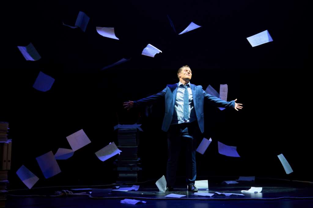 Henkilö seisoo lavalla kädet levällään, ilmassa leijuu papereita, kuvituskuva Nokia-näytelmästä.